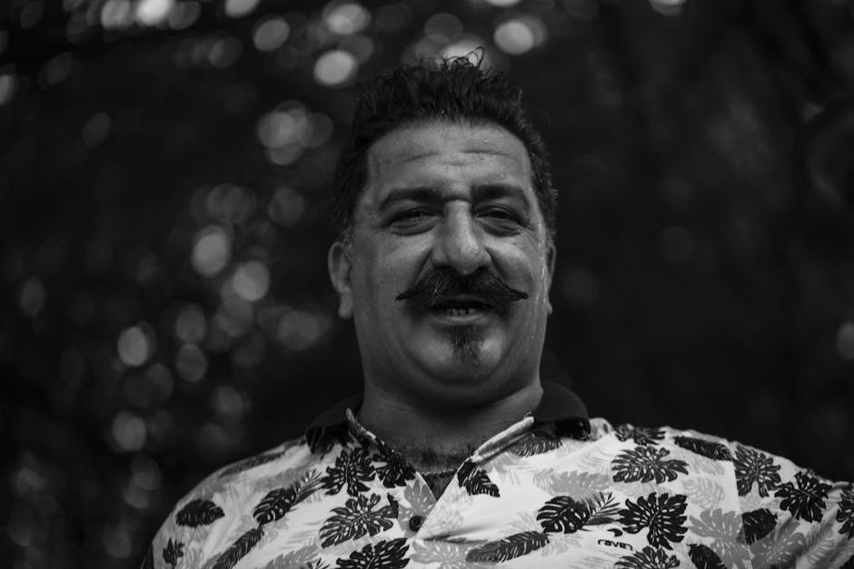 A monochrome portrait of a smiling man with a mustache in a floral shirt.