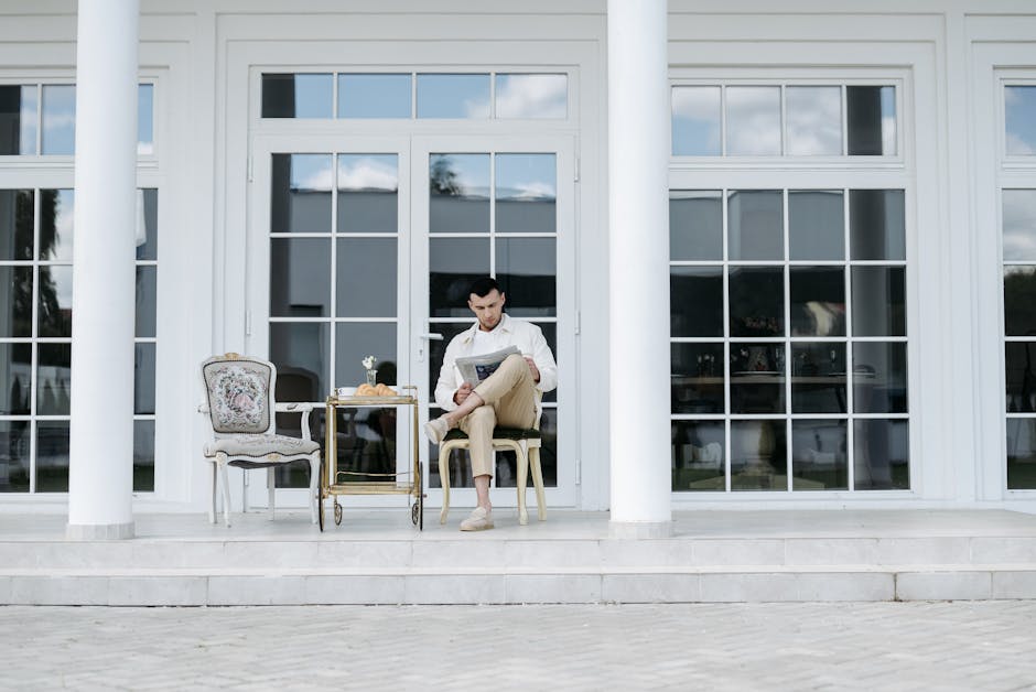 Man sitting on a porch reading a newspaper, enjoying leisure time with a cart beside him.