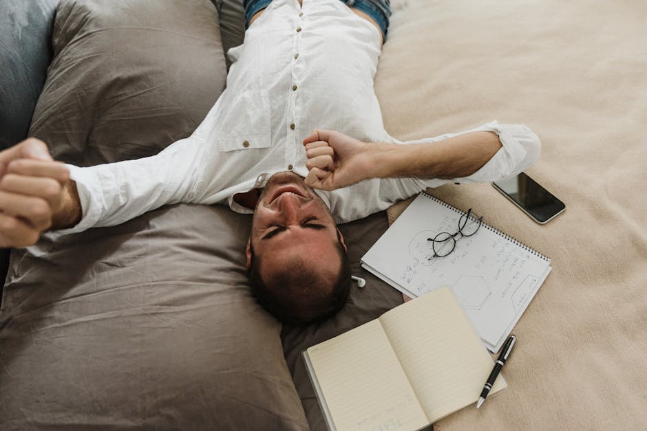 A tired man stretches on a bed surrounded by a notebook, pen, and phone, depicting work exhaustion.