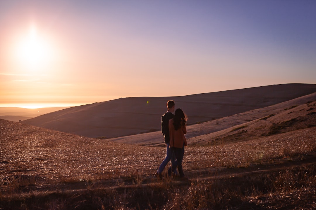 a man and woman kissing on a dirt road in front of a sunset