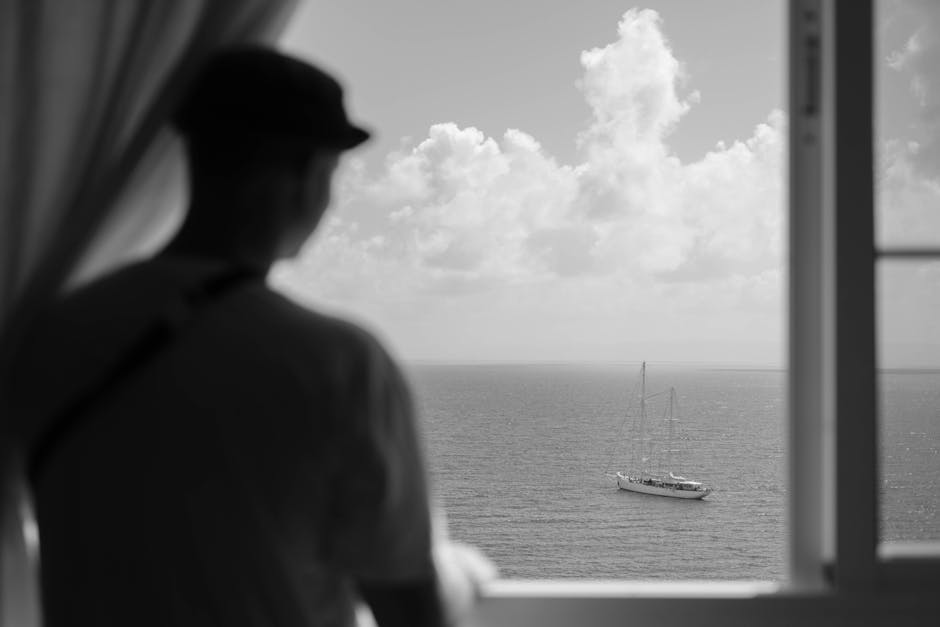 A contemplative man looks out at a sailboat on the sea, captured in black and white.