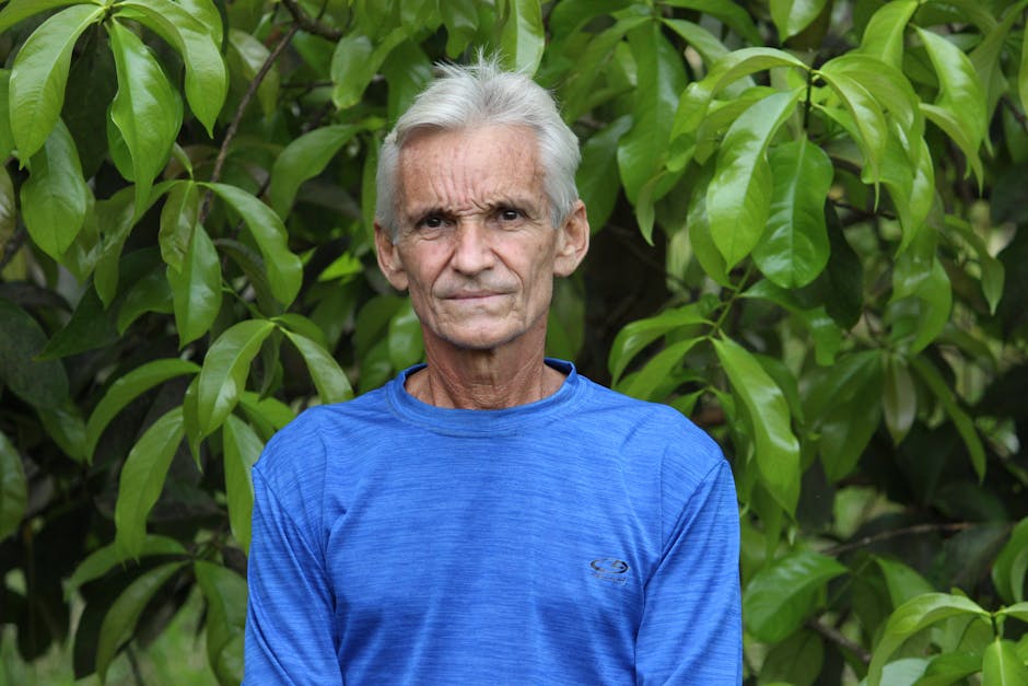 A senior man in a blue shirt stands outdoors against a lush green foliage background. Outdoor portrait.