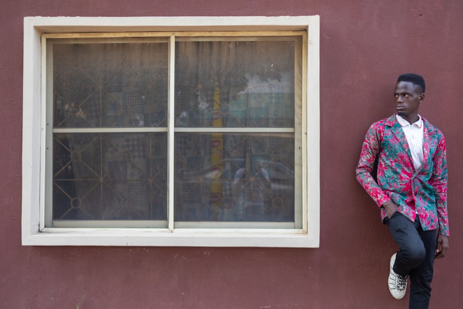 A man in a colorful blazer leaning against a rustic wall near a window.