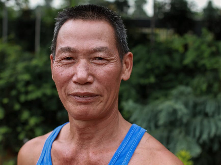 Smiling middle-aged man in blue tank top posing outdoors with greenery background.