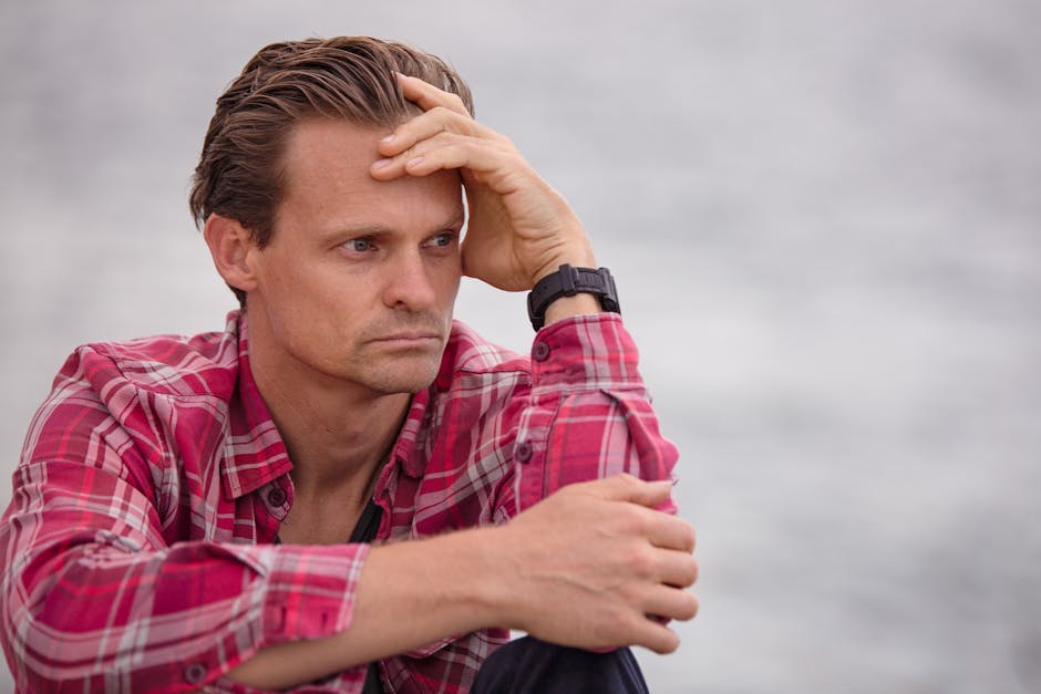 Adult male in plaid shirt sitting outdoors, deep in thought, with casual and relaxed demeanor.