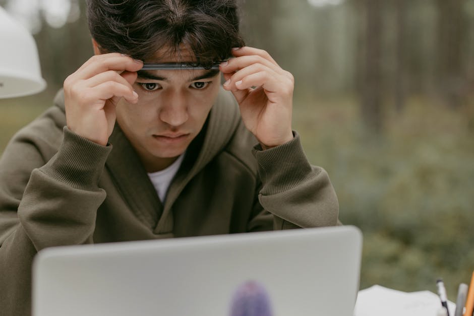 A young man in a green jacket concentrating on a laptop screen in an outdoor setting.