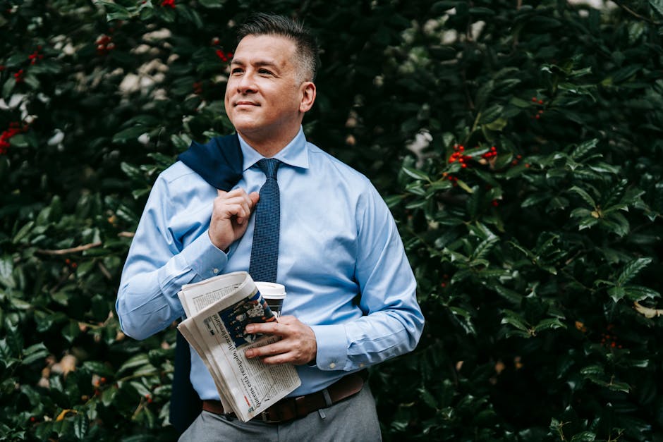 A professional man in formal wear holding a newspaper confidently outdoors, surrounded by greenery.